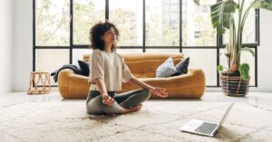 Young woman meditating in her living room in front of a laptop.