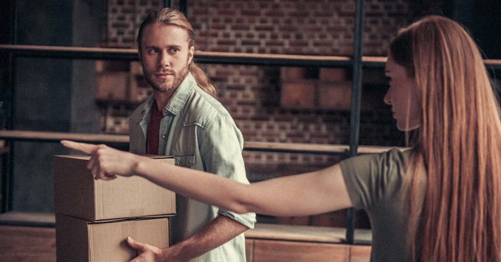 Woman pointing toward door as partner holds moving boxes during relationship separation