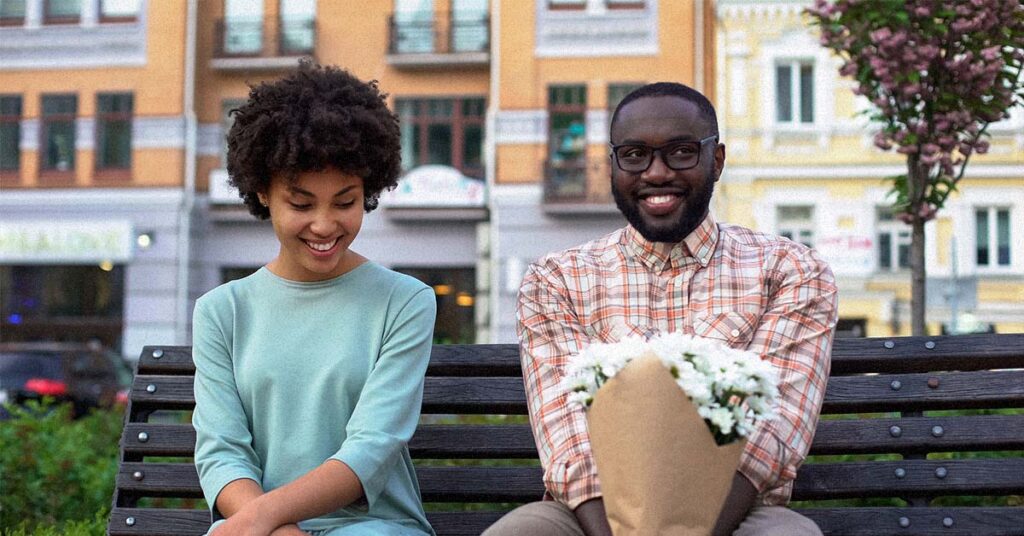 Couple sharing a joyful moment on park bench, man holding flowers while woman smiles shyly